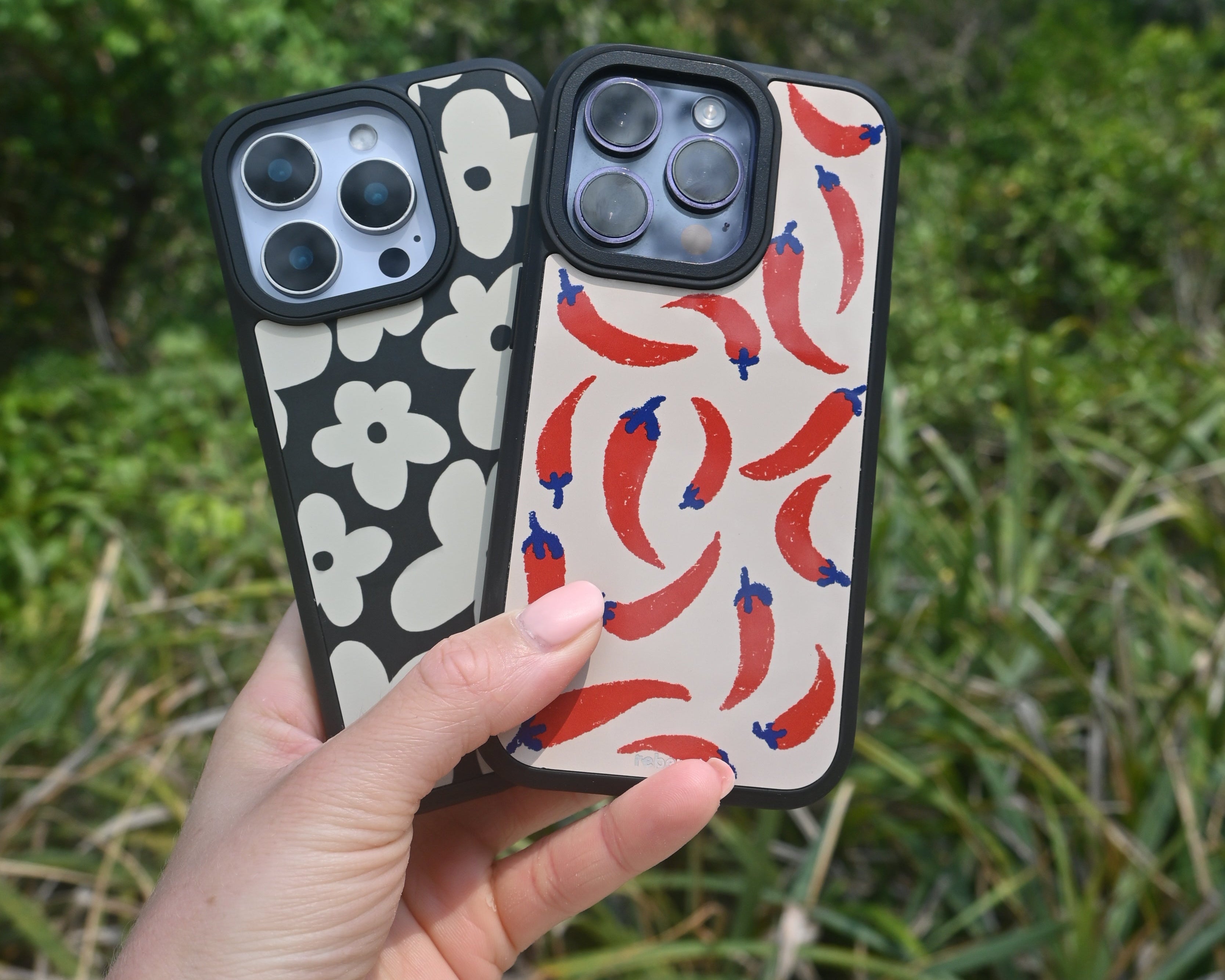 Hand holding two phone cases with floral and fish patterns against a natural background